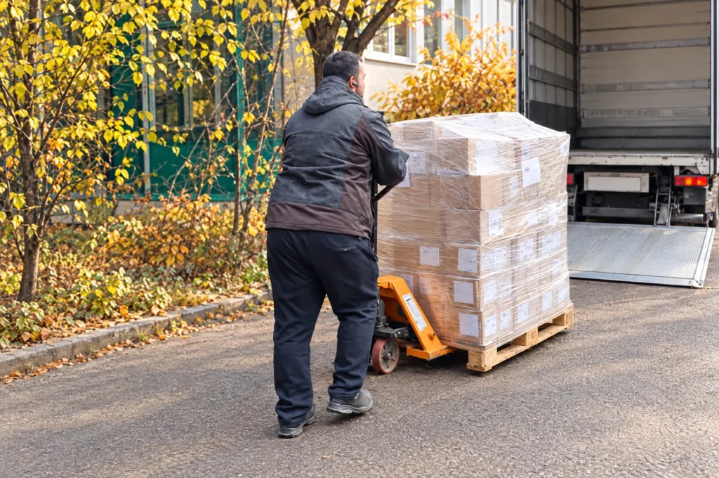 A worker pulling a wrapped pallet of labelled cartons on a pallet jack towards a truck