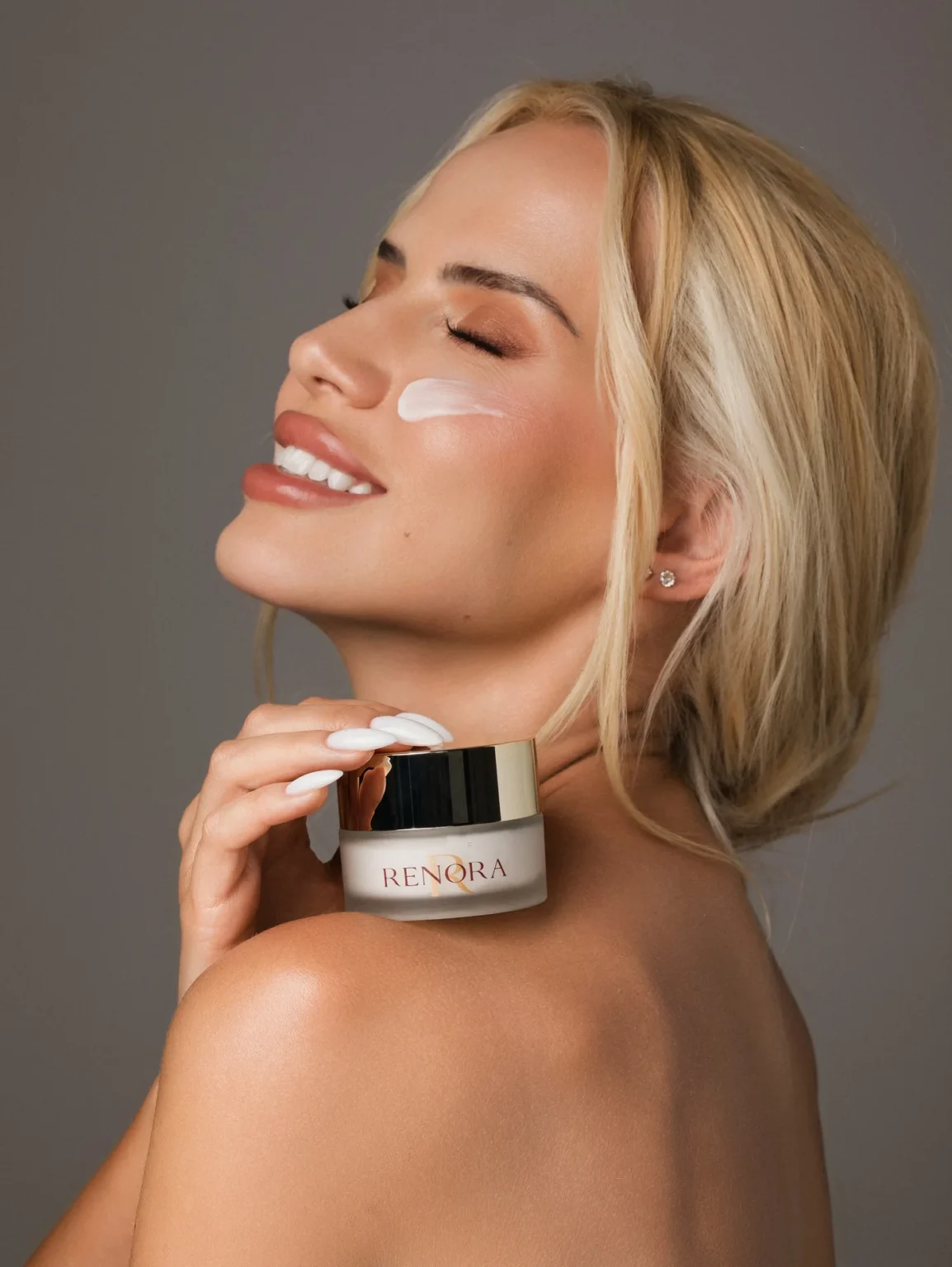 A blonde model with cream on her cheek holding a RENORA cream jar